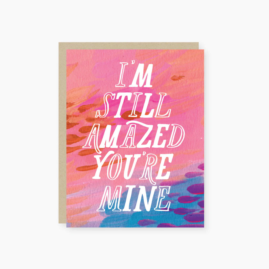 A greeting card with a colorful background and the text 'I'm Still Amazed You're Mine' in bold letters. The card also has a matching envelope.