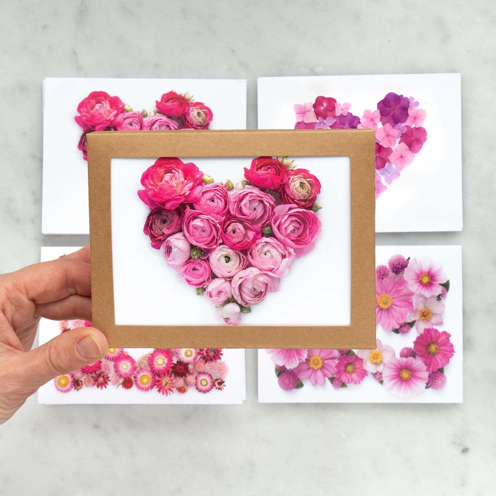 Heart-shaped arrangement of pink flowers in a wooden frame held by a hand on a light gray background.