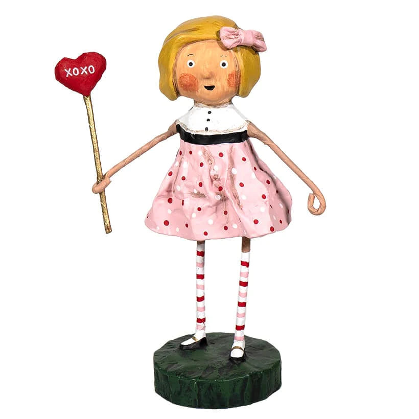 A whimsical figurine of a blonde girl holding a red heart-shaped sign that reads "XOXO." She wears a pink polka-dot dress with a white collar and black waistband, red and white striped socks, and black shoes, standing on a dark green base.
