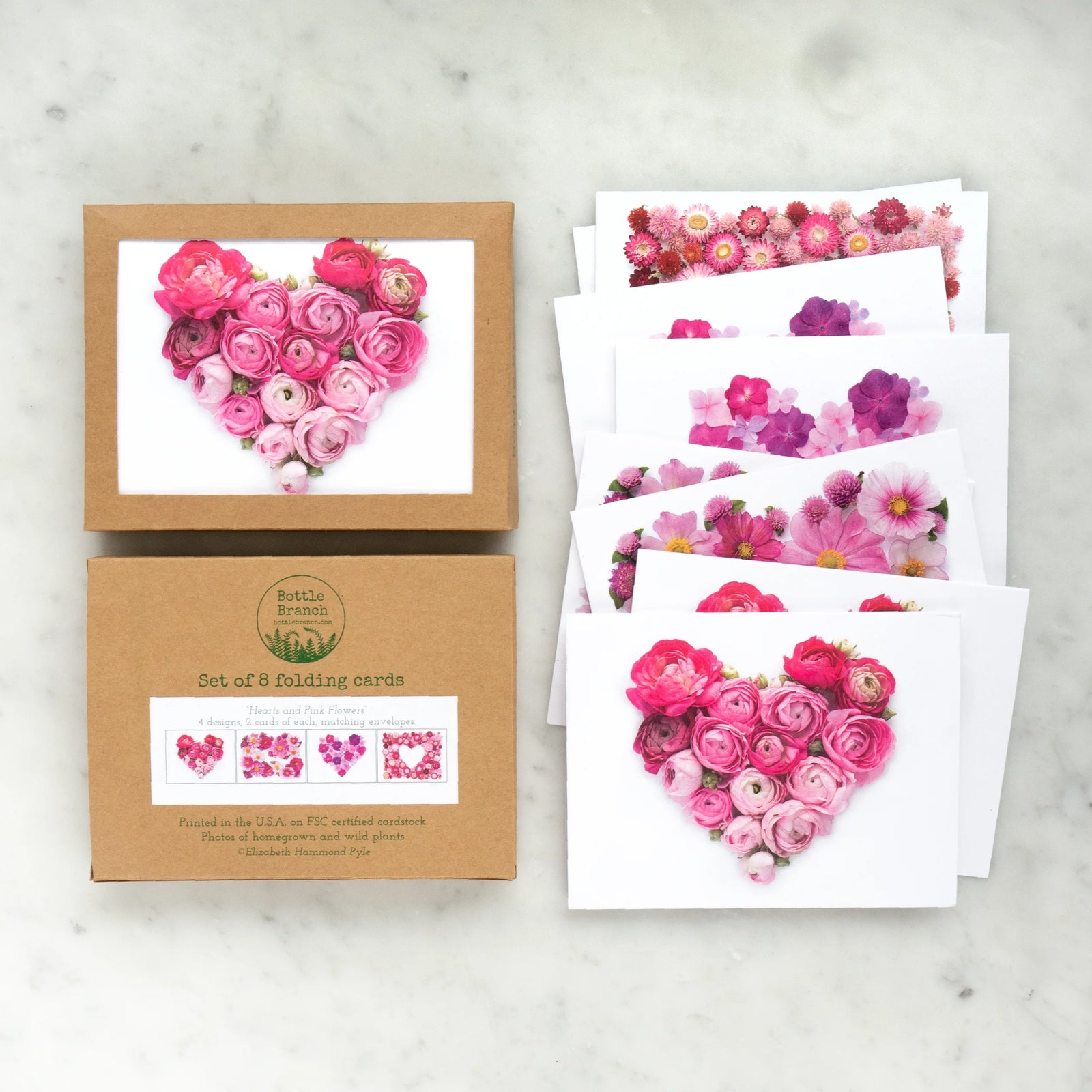 Heart-shaped arrangement of pink flowers in a wooden frame held by a hand on a light gray background.