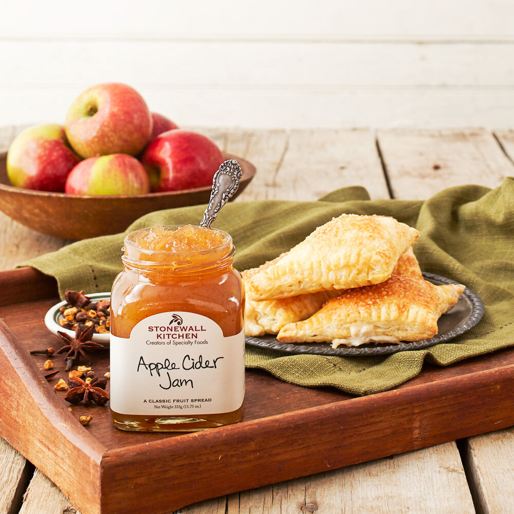 A open jar of Stonewall Kitchen Apple Cider Jam with a silver serving spoon inside sits on a wooden tray next to a plate of flaky pastries, with a small bowl of spices. In the background is a wooden bowl with freshly picked red apples.