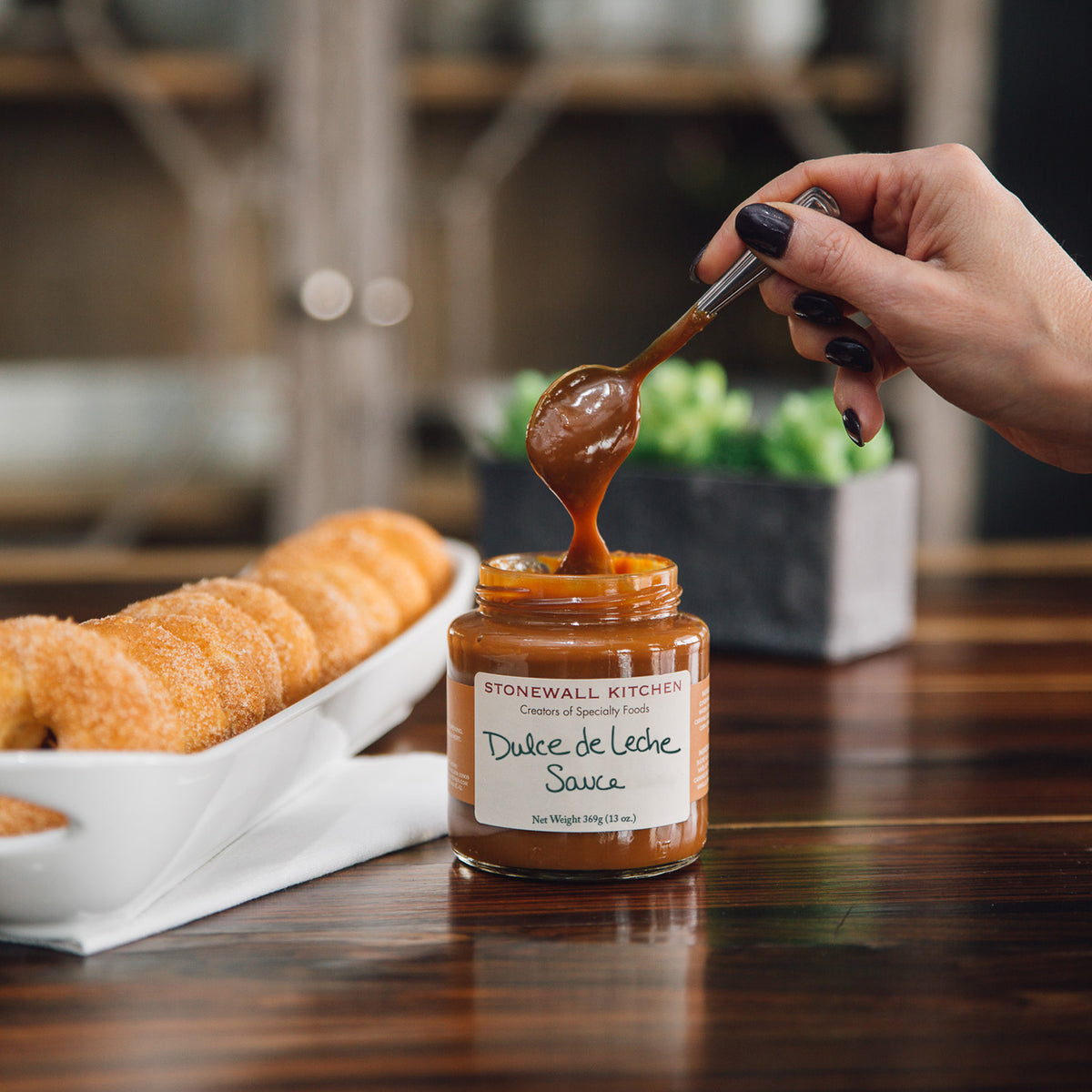 Sugar doughnuts in a tray and hand holding spoon of Stonewall Kitchen dulce de leche sauce dripping in to jar