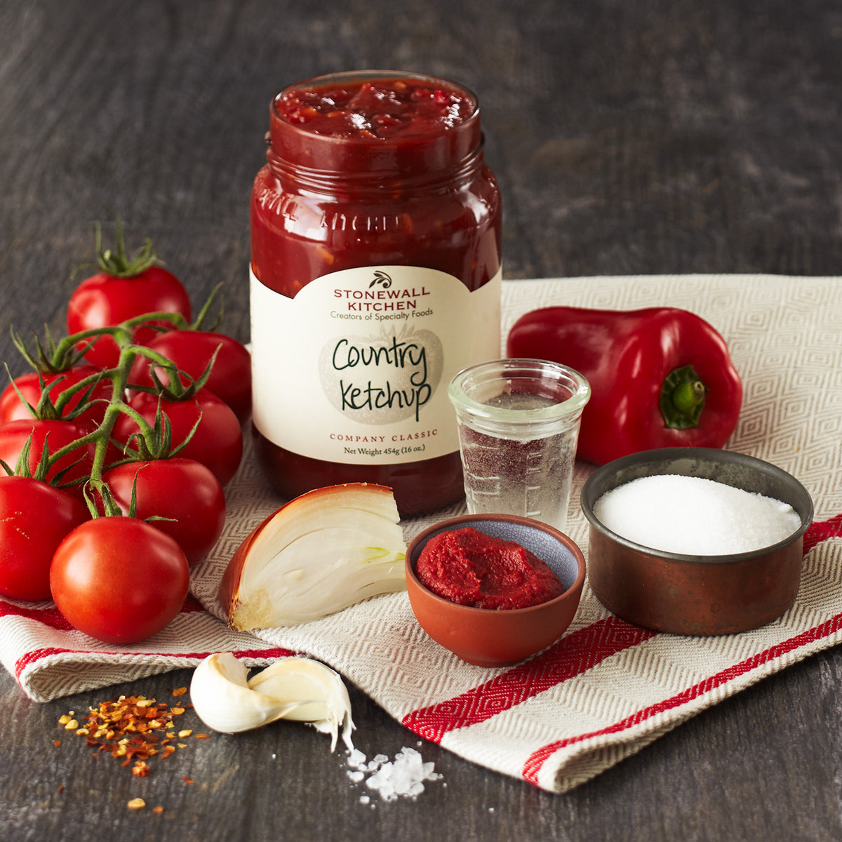 Photo Of A Jar Of Stonewall Kitchen Country Ketchup Surrounded By Ingredients Used In Making It Including Tomatoes Garlic Red Pepper Flakes Salt Onion, Sugar, Red Pepper, and Oil